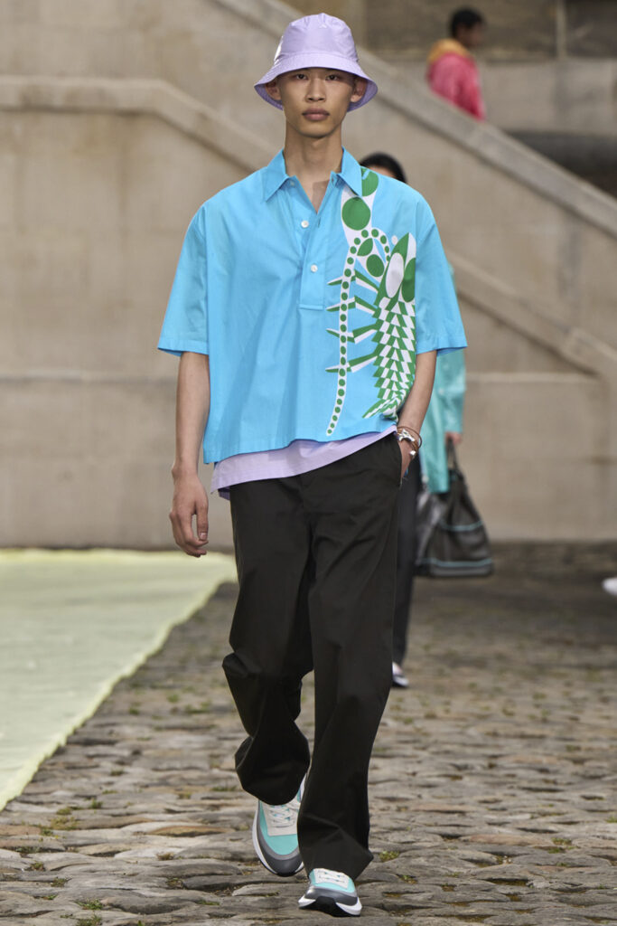 Model in head to toe Hermes with bright blue shirt and lavender hat with black pants and lighter shoes